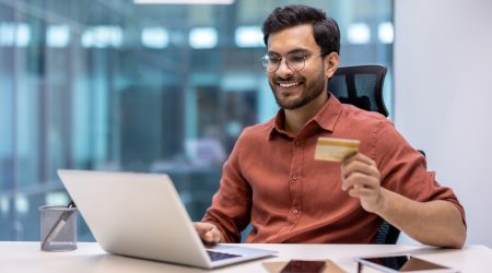 Smiling man in office holding credit card while shopping online and using laptop for secure